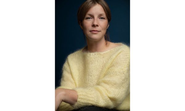 A woman wearing a yellow sweater sits in front of a dark background.