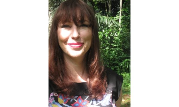 A woman in a colourful shirt is smiling and standing in a lush garden.