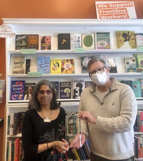 A woman and a man, both wearing masks, are standing in front of a bookshelf and holding a clear plaque.