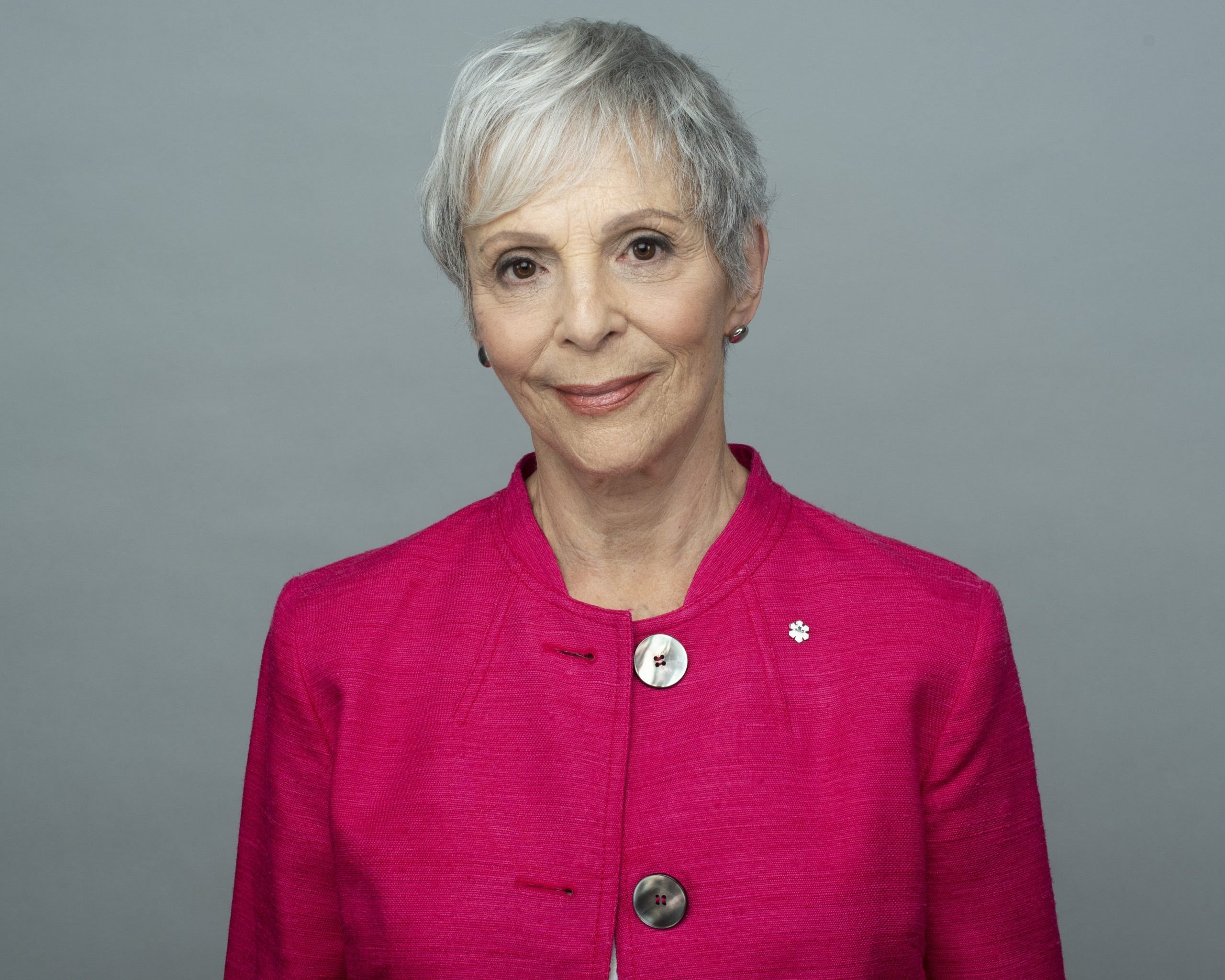 A woman with short silver hair is smiling and wearing a pink suit.