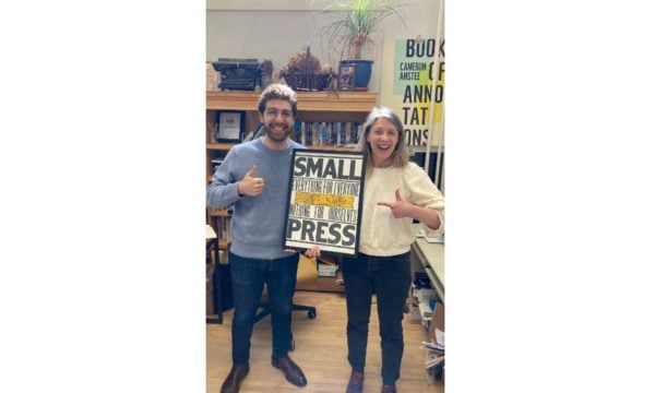 A man wearing jeans and a blue sweater and a woman wearing jeans and a cream-coloured sweater are smiling and holding a sign that says "small press."