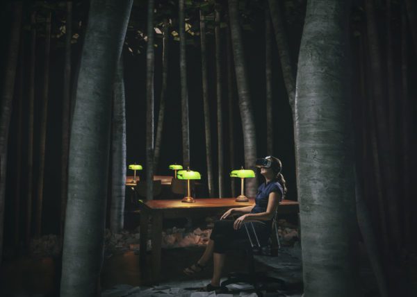A person is seated at a wooden table with green lamps, wearing a virtual reality headset and surrounded by tall grey tree trunks