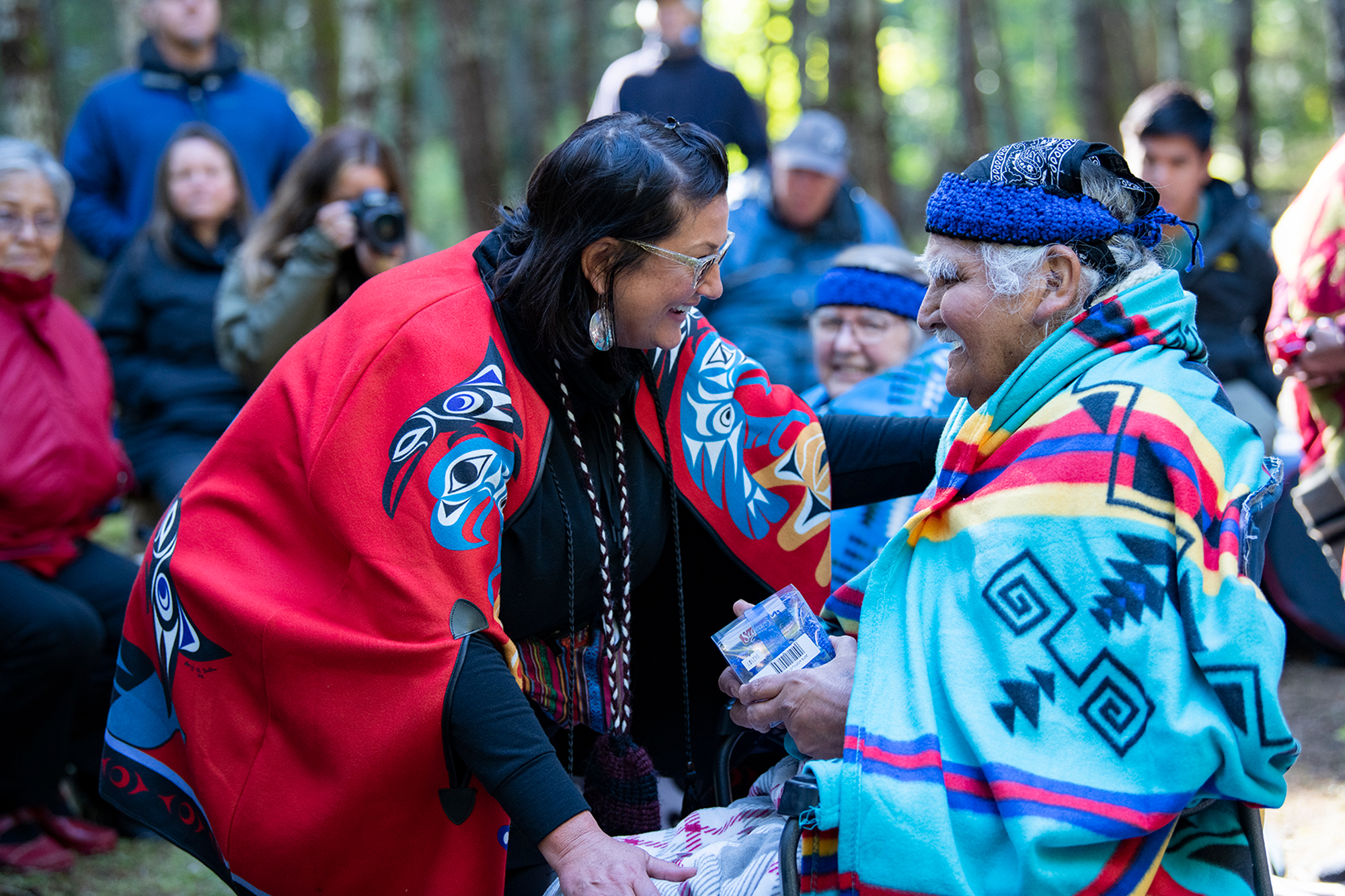 A seated Cowichan elder is greeted at a ceremony in Nanaimo while a small group looks on in the background.