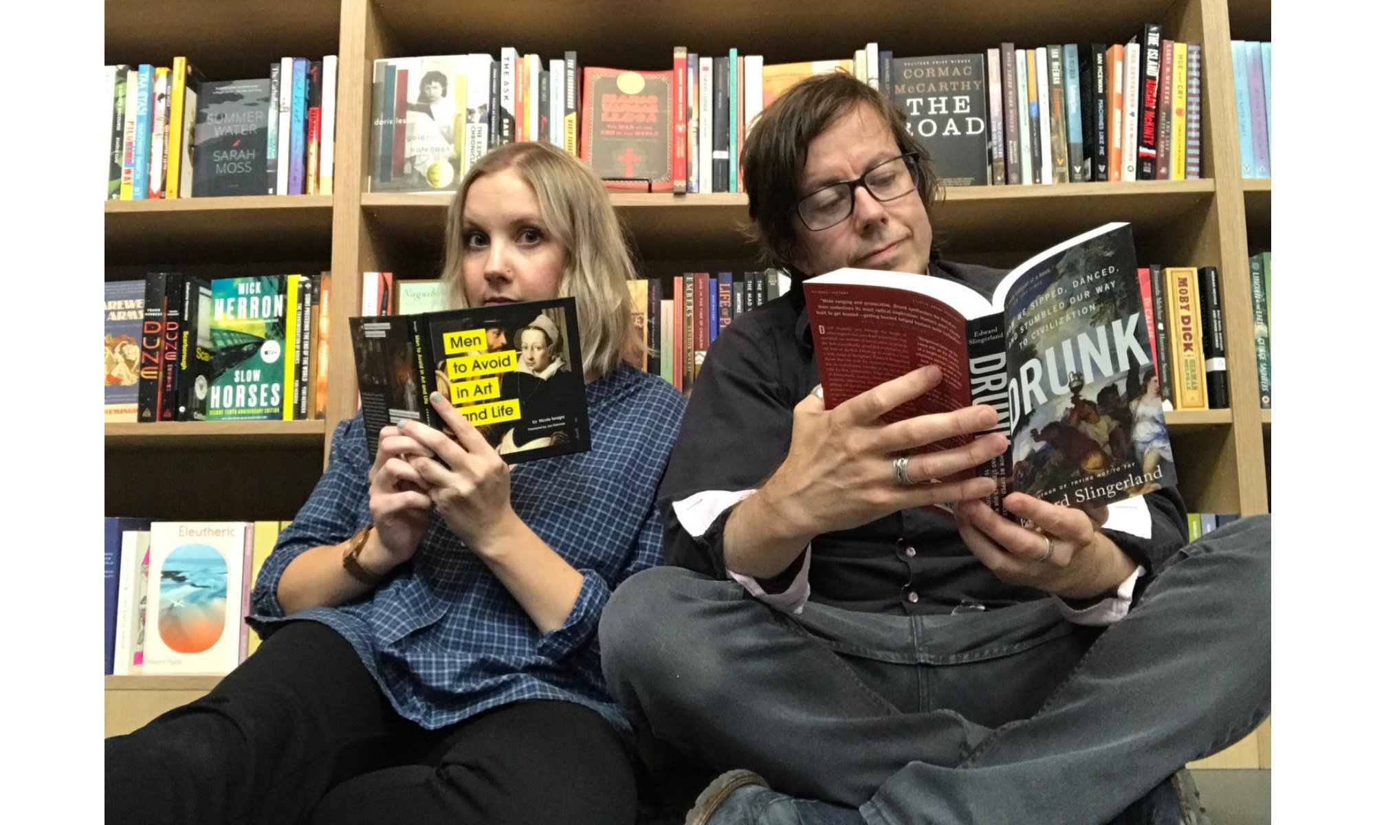 A man and a woman sit in front of a bookshelf. She is reading a book called "Men to Avoid in Life and Art" and he is reading a book called "Drunk."