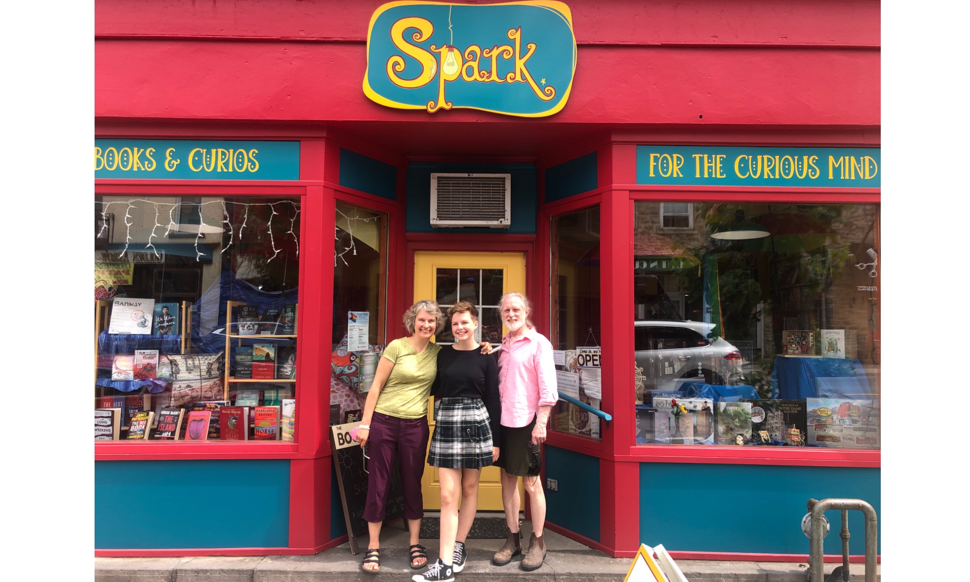 Three people stand in front of a red storefront. The sign says "Spark" in yellow letters on a turquoise background.