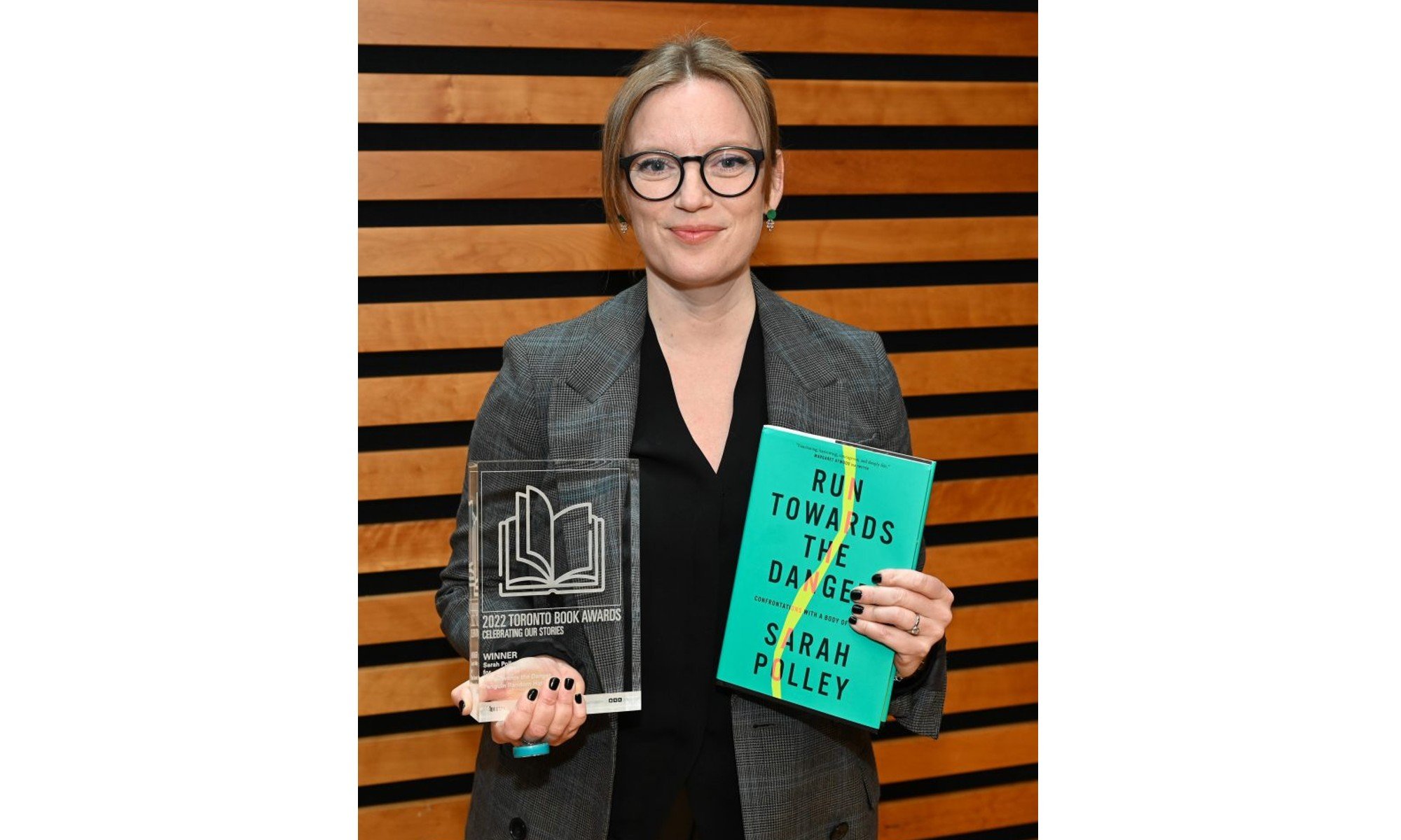 A woman blonde woman wearing glasses holds a book and an award made of glass.
